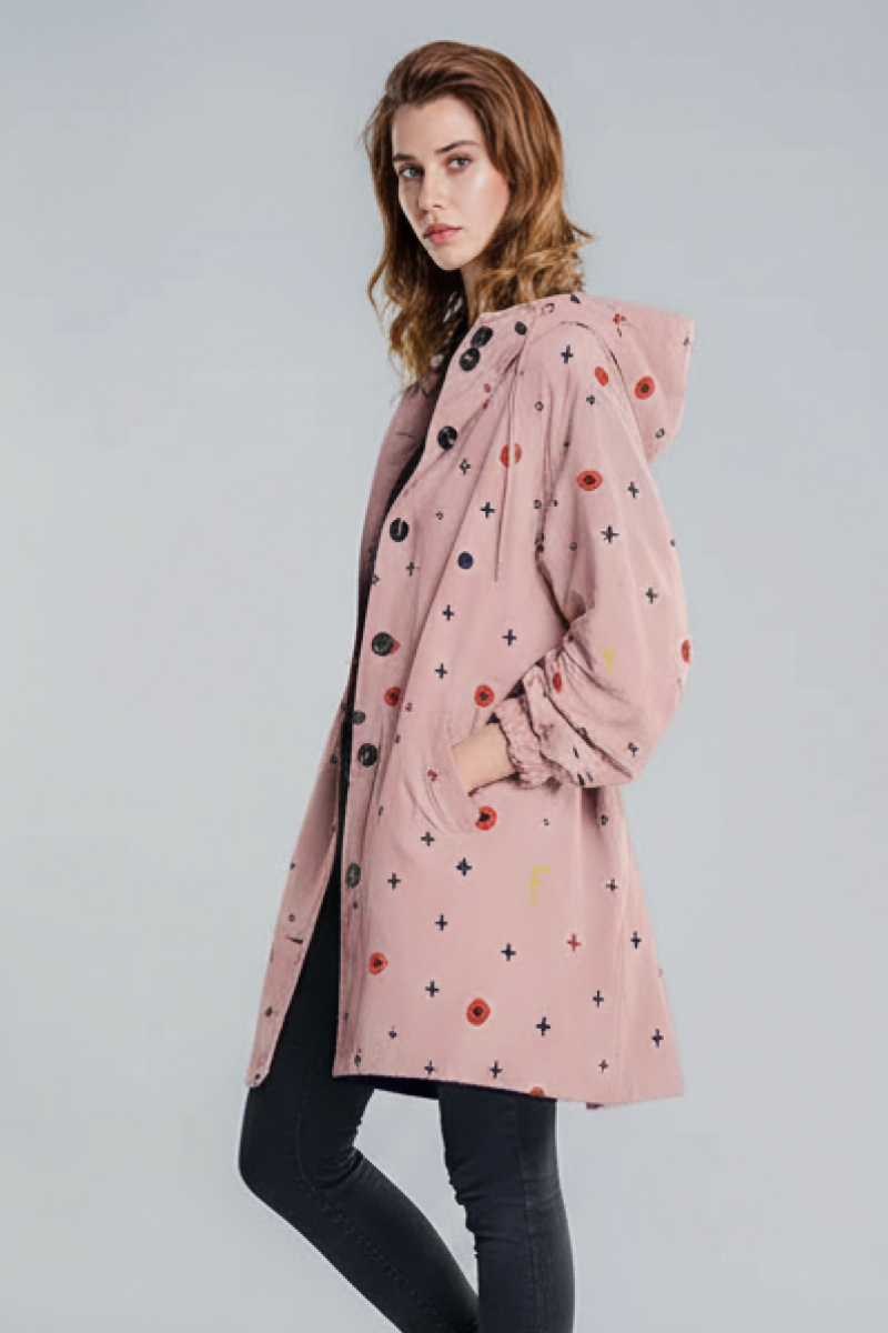 A woman in profile wears the looselyboho DOT & BLOOM WINDBREAKER—light pink, water-resistant, with black buttons and a pattern of small black crosses and red dots—paired with dark skinny jeans against a plain background.