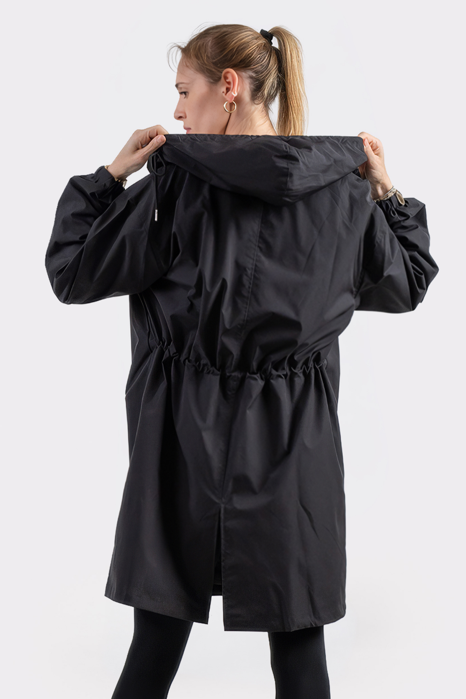 A woman with light hair in a ponytail stands with her back to the camera, wearing the Icon Windbreaker Rain Jacket and black leggings. She holds the jacket’s hood with both hands.