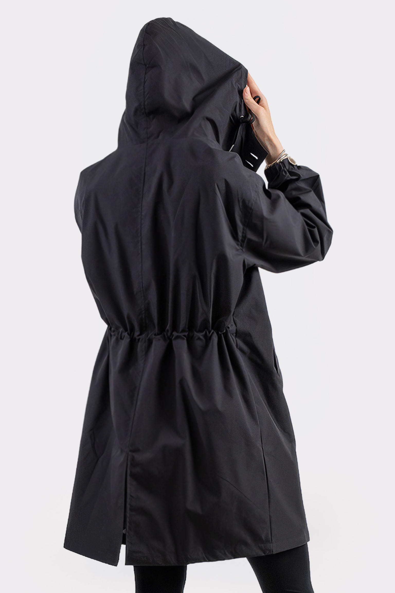 Someone is seen from behind wearing the Icon Windbreaker Rain Jacket—a long black raincoat with a hood, cinched waist, and back hem slit. One hand holds the hood. The background is light and plain.