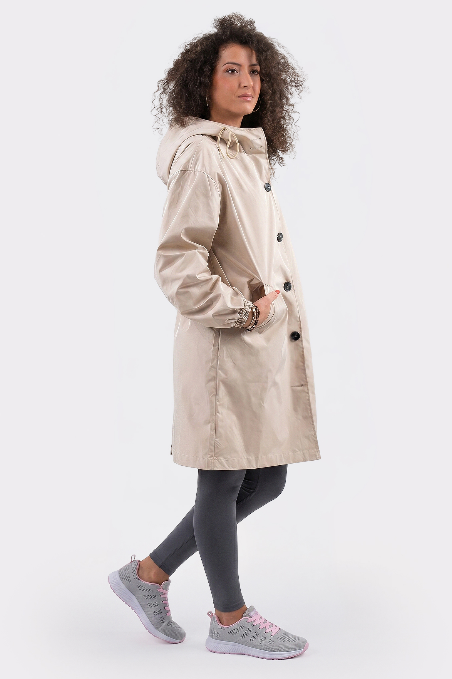 A person with curly hair stands sideways against a plain white background, wearing the Icon Windbreaker Rain Jacket in light beige, paired with dark gray leggings and gray sneakers featuring light pink accents.