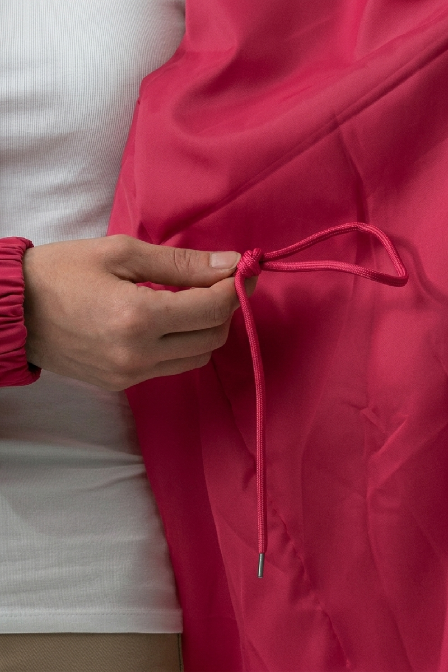 A person adjusts the drawstring of an Icon Windbreaker Rain Jacket in bright pink while wearing a white top.