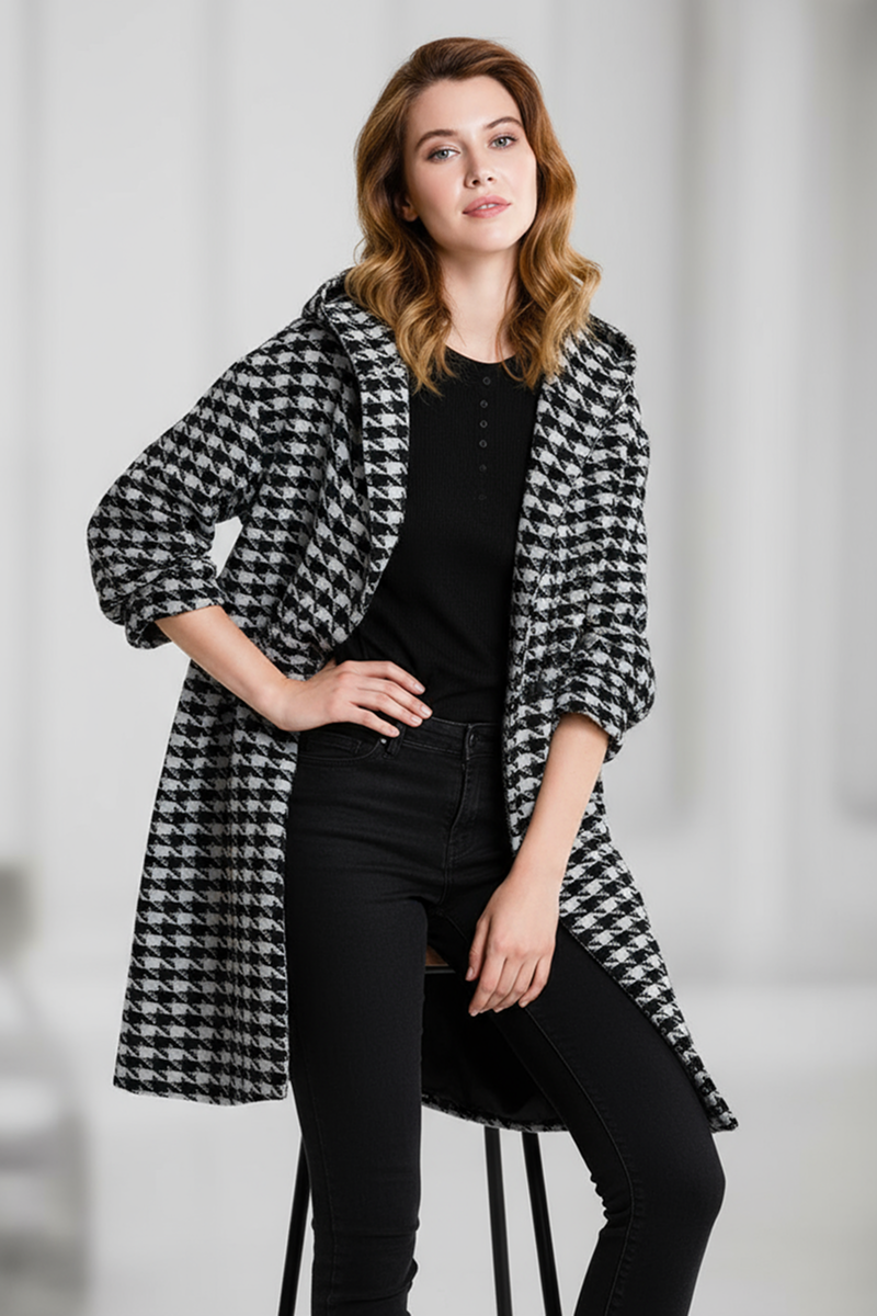 A woman with shoulder-length wavy hair stands indoors wearing the looselyboho HOUNDSTOOTH WINDBREAKER over a black top and jeans, one hand on her hip and the other on her thigh.
