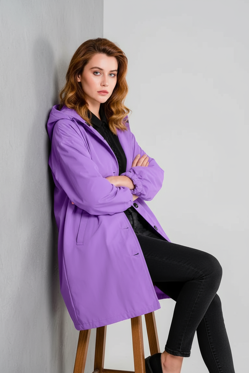 A woman with light skin and brown hair wears the looselyboho SOFT LAVENDER WINDBREAKER with an adjustable hood and black pants, sitting on a stool against a light wall, looking straight ahead with a neutral expression.