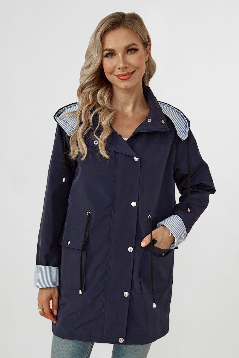 A woman with blonde hair, wearing a navy blue waterproof jacket with silver buttons and drawstrings, smiles at the camera. The Lightweight Windbreaker showcases light blue striped lining visible at the cuffs and hood. She has one hand in her pocket and stands against a plain white background.
Product Name: STRIPED NAVY SHADOW WINDBREAKER JACKET
Brand Name: looselyboho