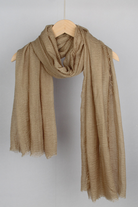 A light brown, textured scarf hangs on a wooden hanger against a plain background. The looselyboho Color-POP Scarf is loosely draped, showcasing its lightweight and soft fabric with a slightly crinkled texture, making it an ideal accessory for all-weather wear.