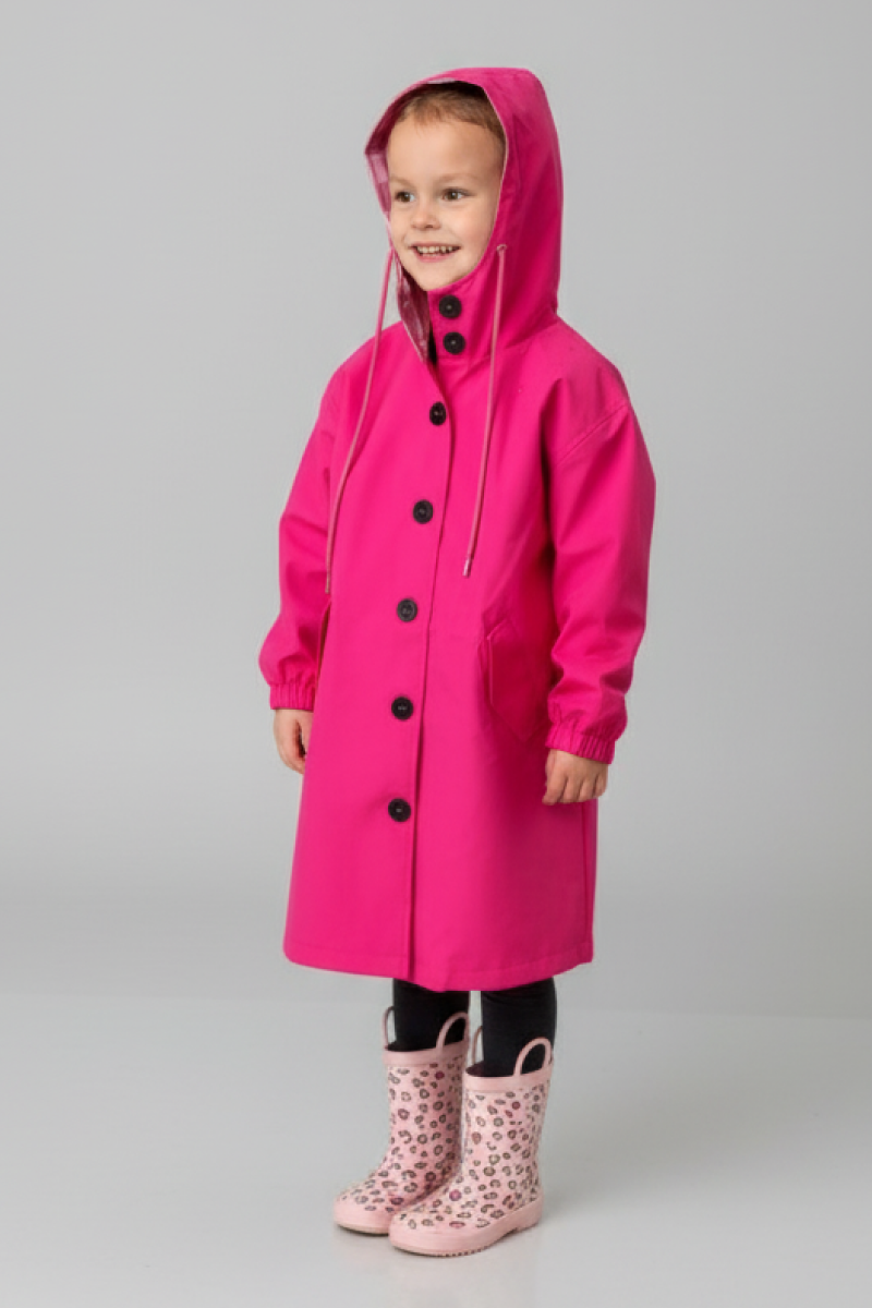 A young child in the looselyboho Bright Berry Kids’ Windbreaker with black buttons and pockets, paired with light pink leopard print rain boots, smiles against a plain light gray background.