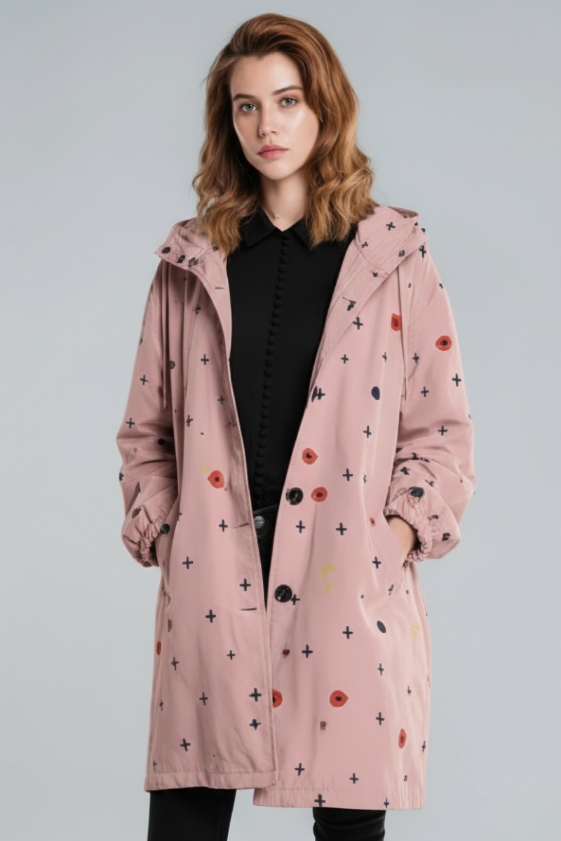 A woman models the looselyboho DOT & BLOOM WINDBREAKER—a light pink, water-resistant jacket with multicolored geometric dots and plus signs—layered over a black button-up and pants, set against a plain gray background.