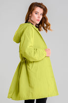 A person with long hair is wearing a SUNLIT YELLOW WINDBREAKER by looselyboho and black pants. They are standing sideways, slightly facing the camera against a gray backdrop, highlighting the windbreaker's water-resistant design.