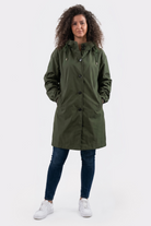 A person with curly hair wears the Icon Windbreaker Rain Jacket, styled in a long olive green design, along with blue jeans and white sneakers. They stand facing forward, hands in pockets, against a plain white background.