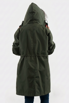 A person in blue jeans is shown from the back wearing the Icon Windbreaker Rain Jacket in olive green with the hood up, adjusting it. The background is plain and light-colored.