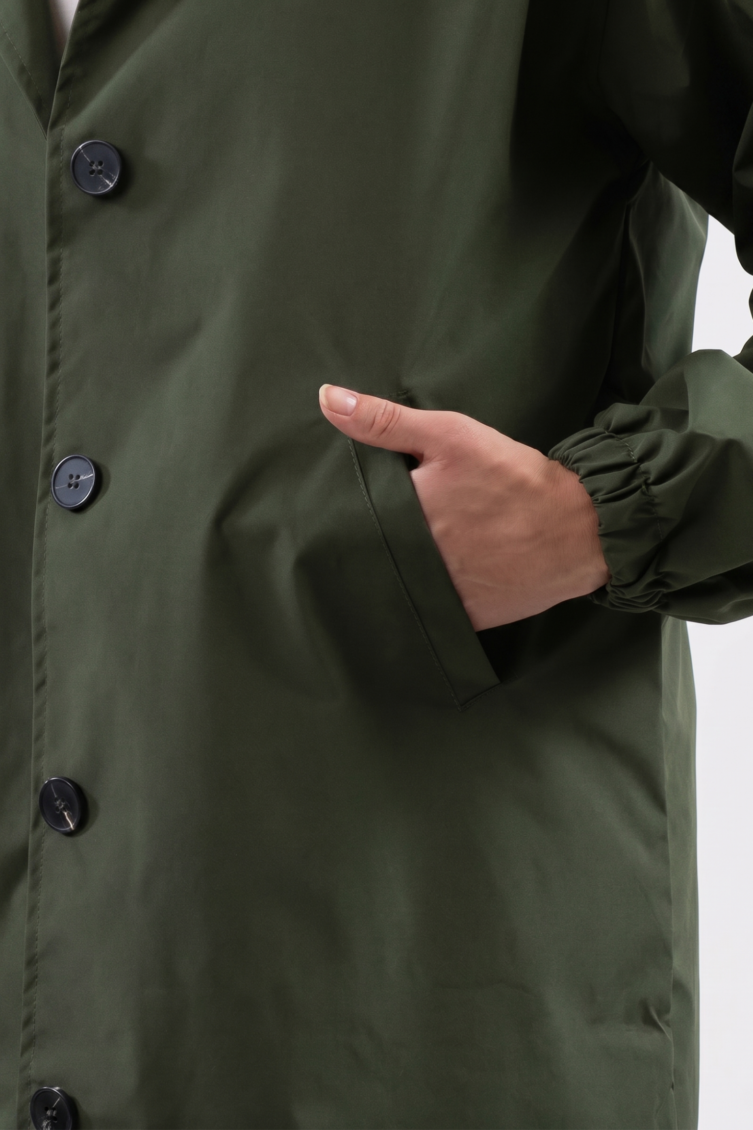 A person wears The Water Resistant Hooded Oversized Windbreaker Rain Jacket in dark green, featuring black buttons, a straight cut, elastic wrists, and stands with their right hand in the pocket against a plain light background.
