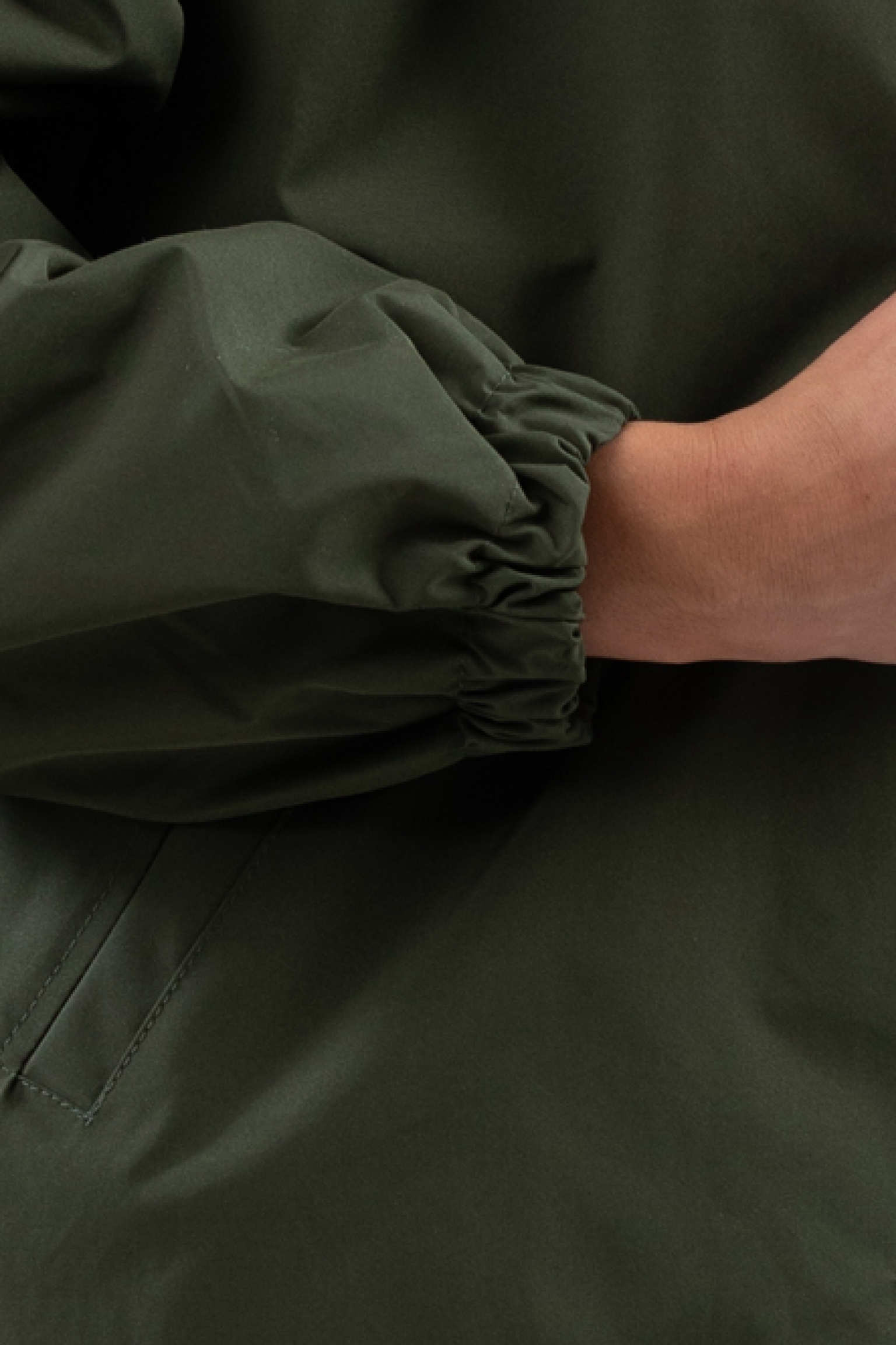 A close-up of a person’s arm wearing The Water Resistant Hooded Oversized Windbreaker Rain Jacket in dark green; their gathered-cuff sleeve and hand are partially visible against a plain background.