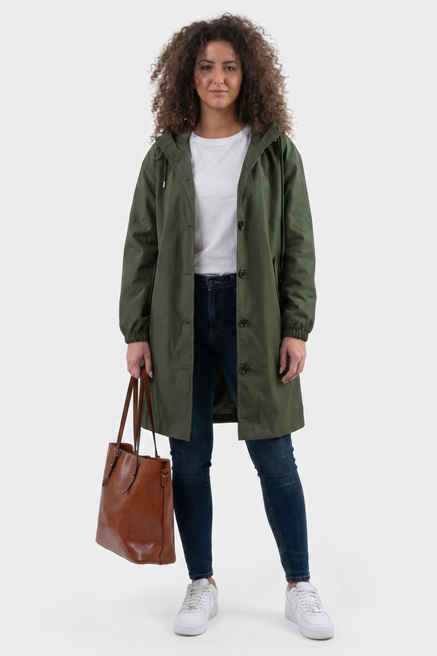A person with curly hair faces forward wearing the Icon Windbreaker Rain Jacket in green, a white t-shirt, blue jeans, white sneakers, and holding a brown leather tote bag in their left hand.