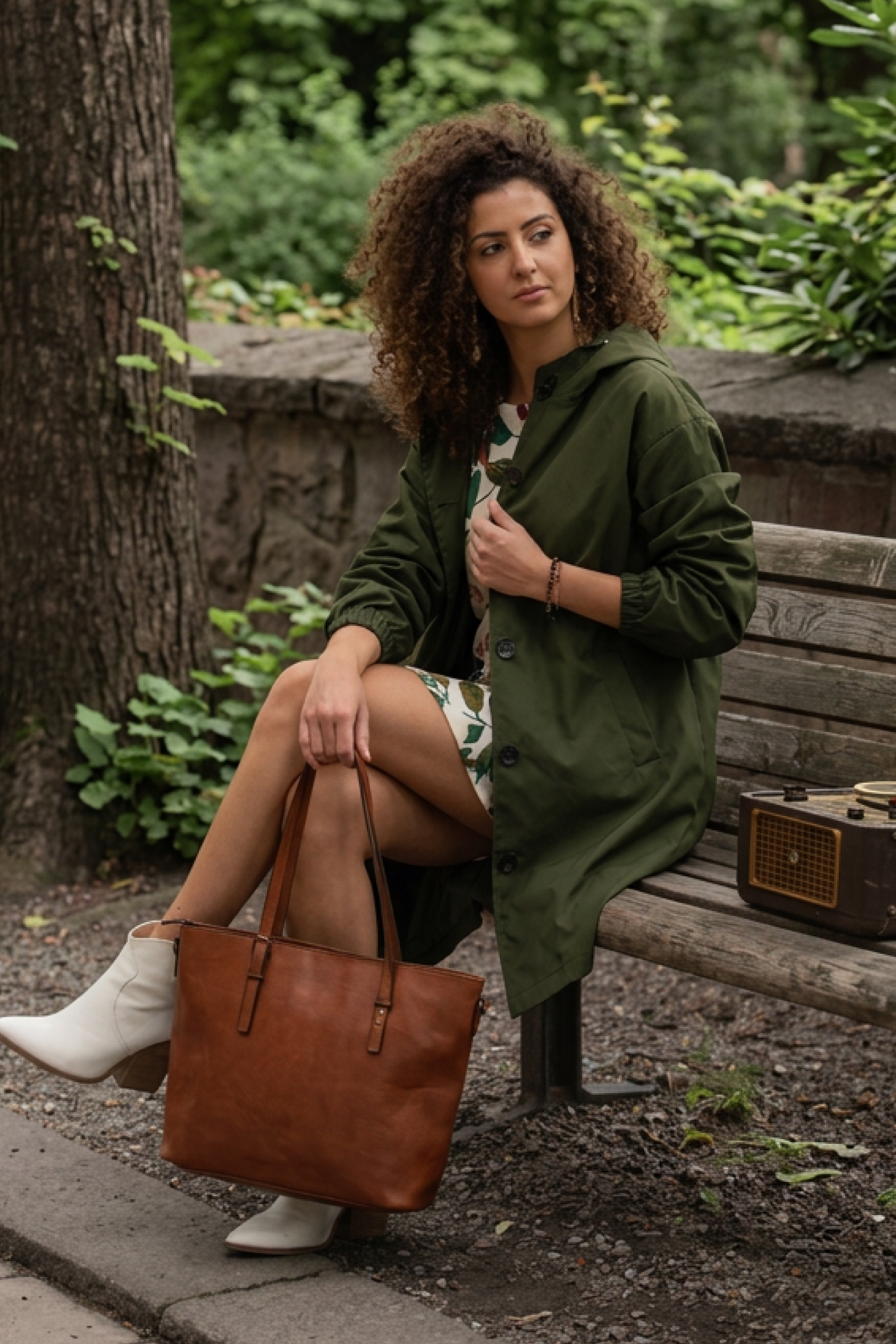 A woman with curly hair sits on a wooden bench outdoors, wearing The Water Resistant Hooded Oversized Windbreaker Rain Jacket, a floral dress, and white boots. She holds a brown leather tote bag, looking to the side beside a vintage radio.