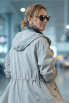 A woman with light hair pulled back stands indoors in a blurred setting, possibly a station, wearing sunglasses and the Icon Windbreaker Rain Jacket in light gray, styled with a patterned scarf.