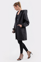 A woman in profile wears the Icon Windbreaker Rain Jacket with black leggings and heeled sandals. Her hair is styled in a ponytail, one hand in her pocket, standing against a plain white background.