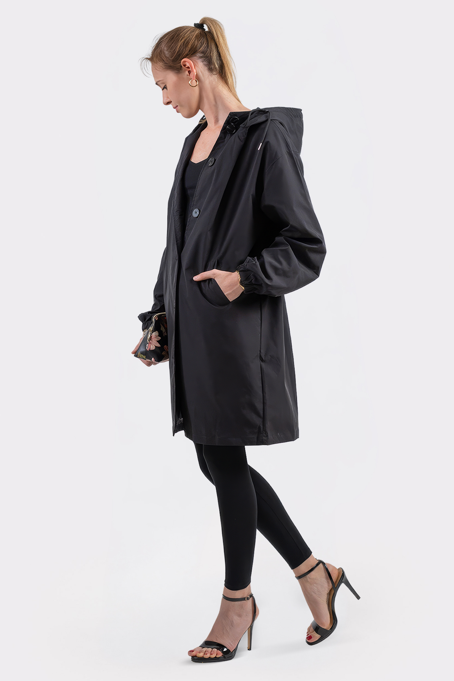 A woman in profile stands against a plain white background, wearing The Water Resistant Hooded Oversized Windbreaker Rain Jacket over black leggings and a top. She holds the pocket and pairs the look with black high-heeled sandals.