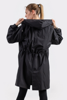 A woman with light hair in a ponytail stands with her back to the camera, wearing the Icon Windbreaker Rain Jacket and black leggings. She holds the jacket’s hood with both hands.