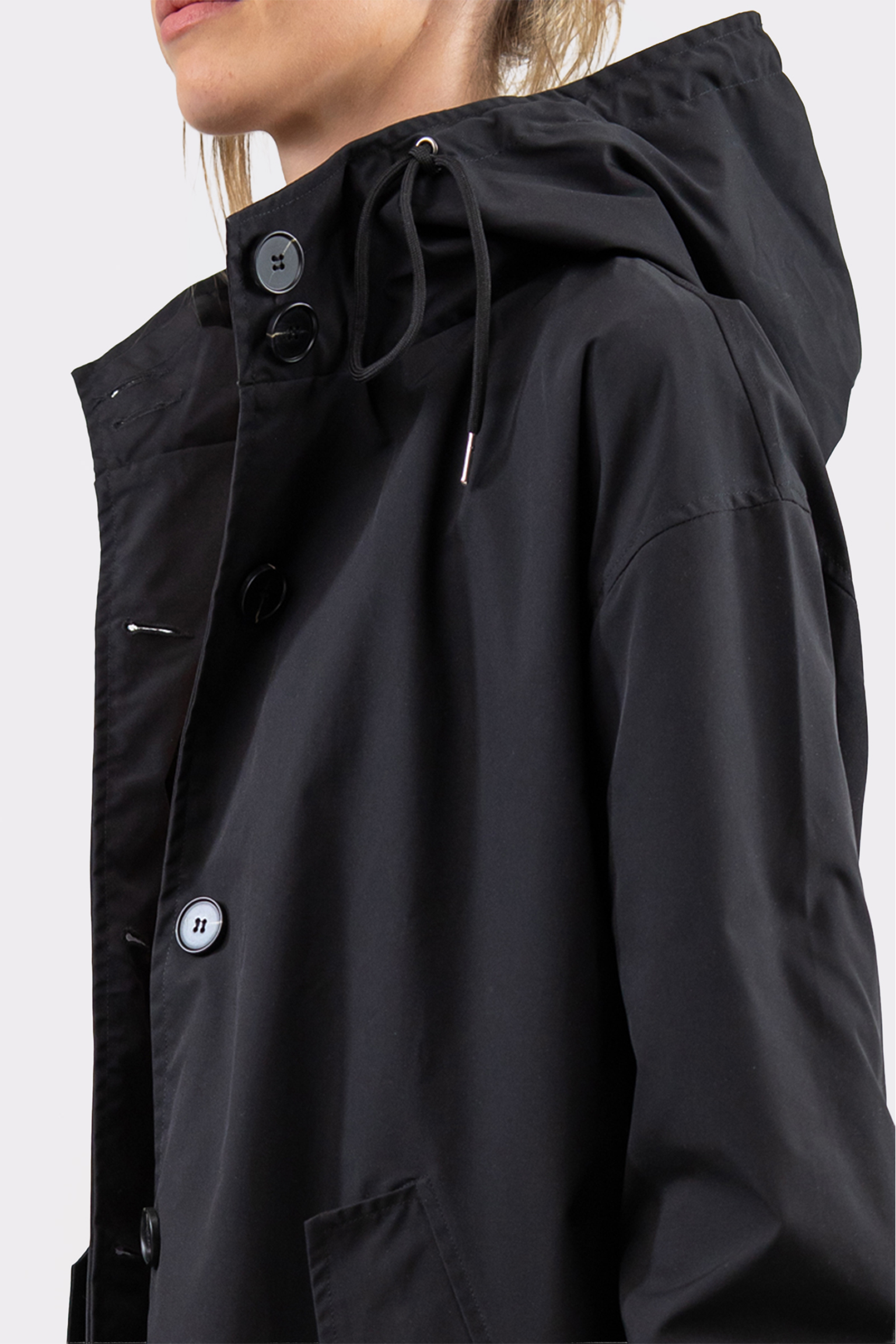 A person wears The Water Resistant Hooded Oversized Windbreaker Rain Jacket, black with large buttons and a drawstring hood, shown in profile against a plain light background.