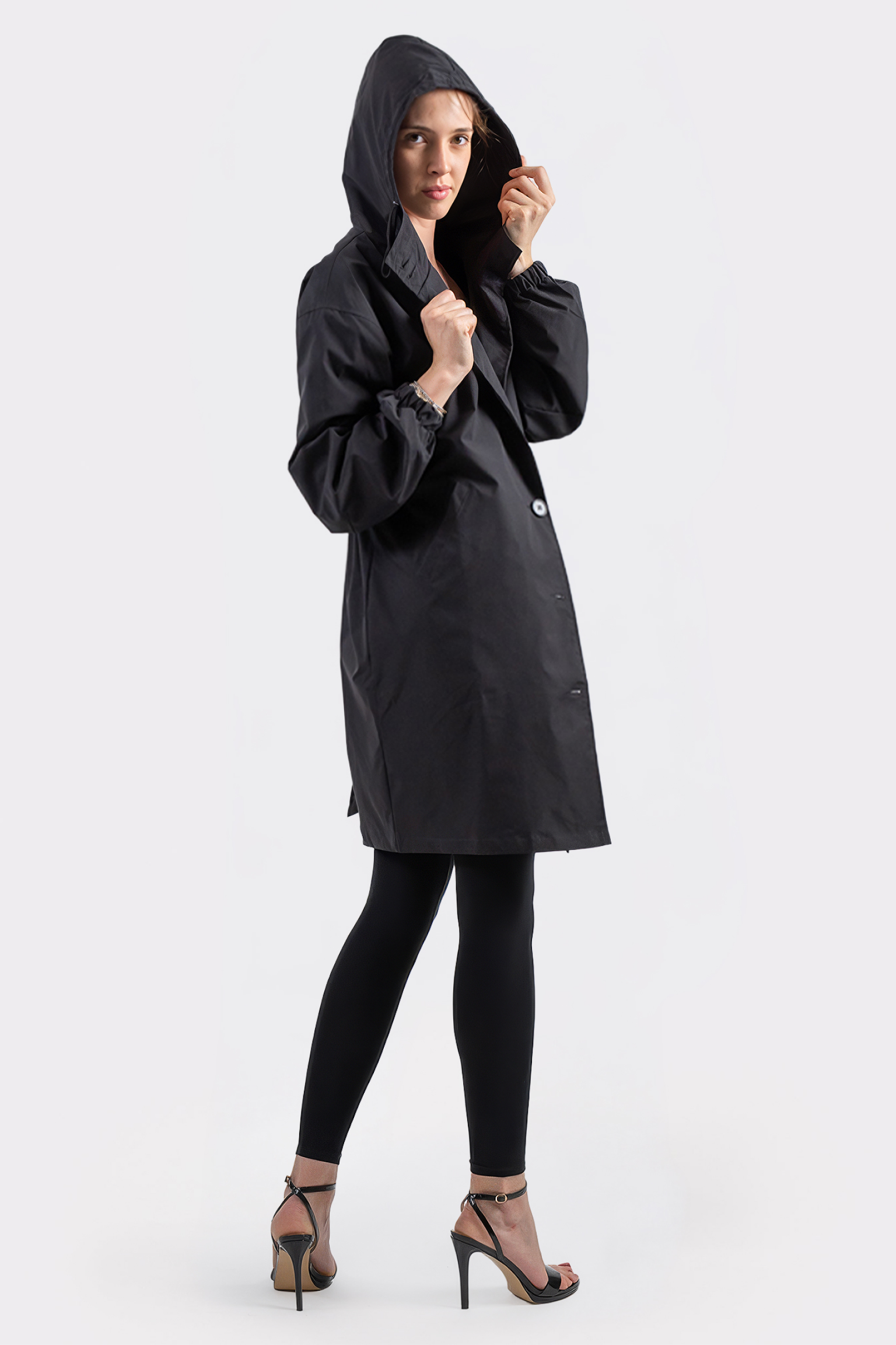 A person models The Water Resistant Hooded Oversized Windbreaker Rain Jacket in black, paired with black leggings and high-heeled sandals, standing slightly sideways against a plain light background.