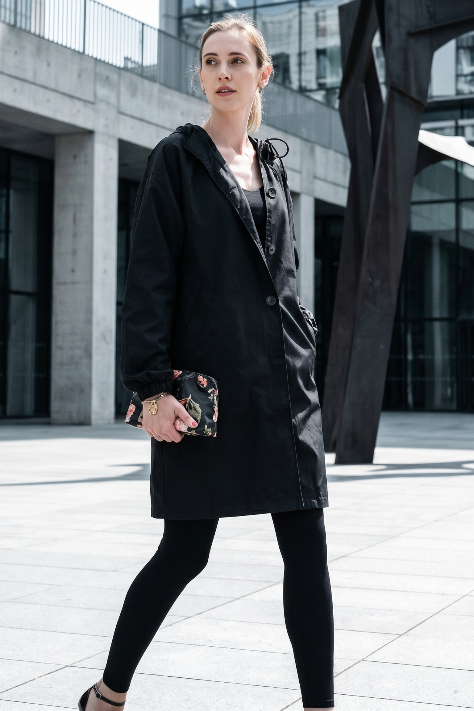 A woman wears the Icon Windbreaker Rain Jacket outdoors in a city with modern buildings, paired with black leggings and holding a patterned clutch as she gazes to the side.
