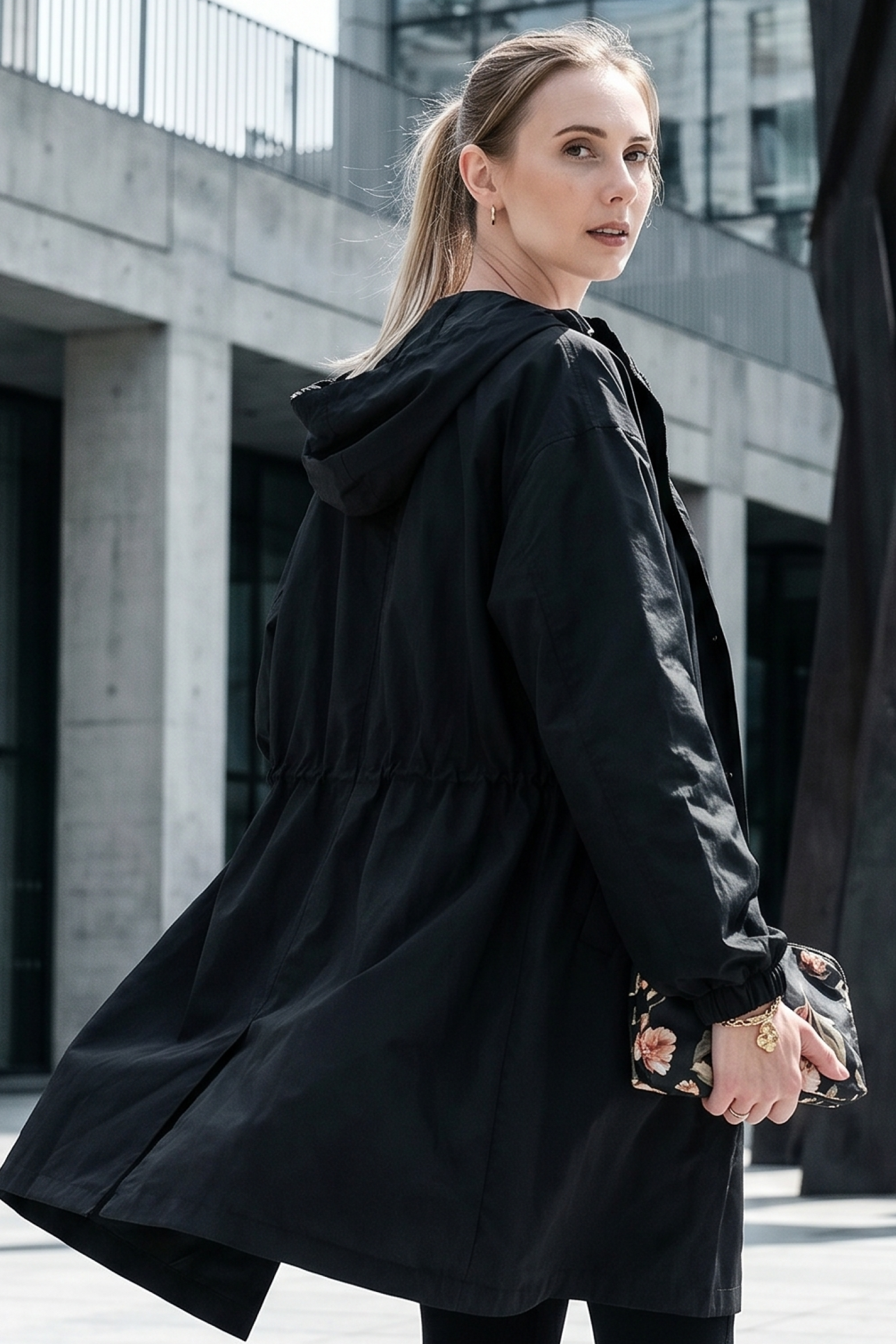 A woman with blonde hair in a ponytail wears The Water Resistant Hooded Oversized Windbreaker Rain Jacket and holds a floral-patterned clutch while standing outdoors near a modern concrete building.