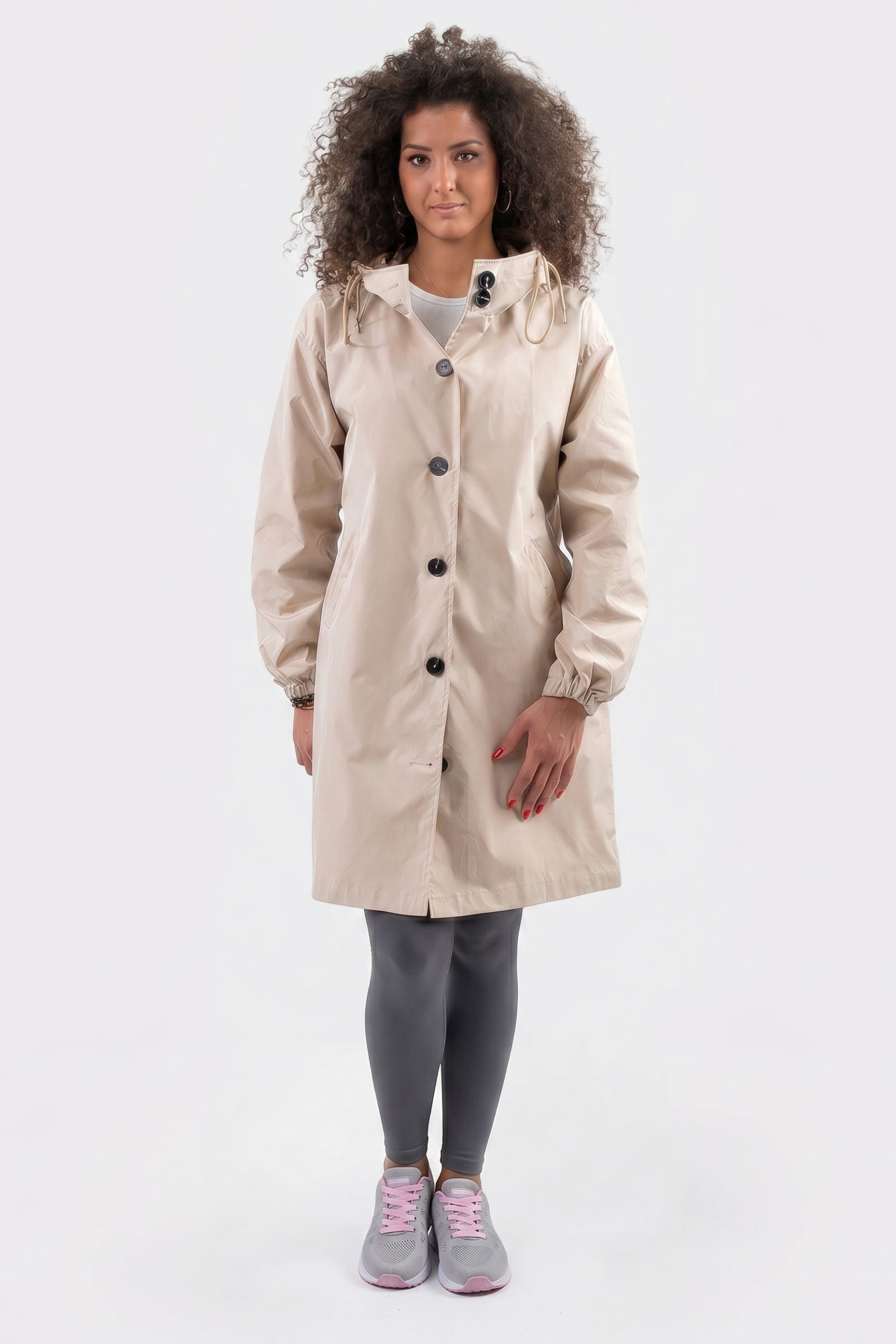 A person with curly hair stands facing forward, wearing The Water Resistant Hooded Oversized Windbreaker Rain Jacket in beige, gray leggings, and gray sneakers with pink laces against a plain white background.