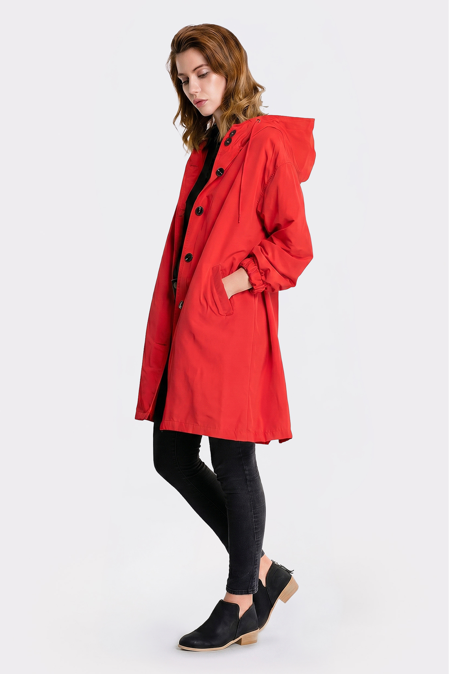 A person stands sideways wearing the Icon Windbreaker Rain Jacket in bright red with black buttons, paired with black skinny jeans and low-heeled black slip-on shoes. Hands are in pockets, set against a plain white background.