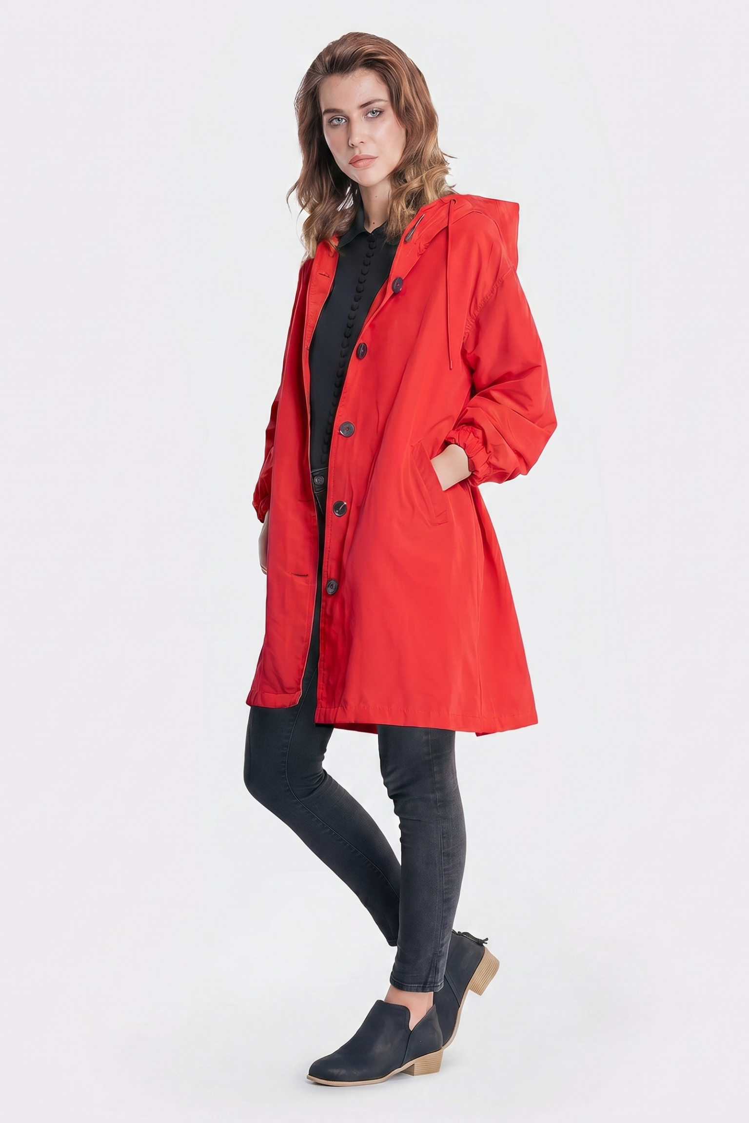 A person models The Water Resistant Hooded Oversized Windbreaker Rain Jacket in bright red, paired with a black shirt, black skinny jeans, and black shoes, standing with one leg slightly bent against a plain white background.