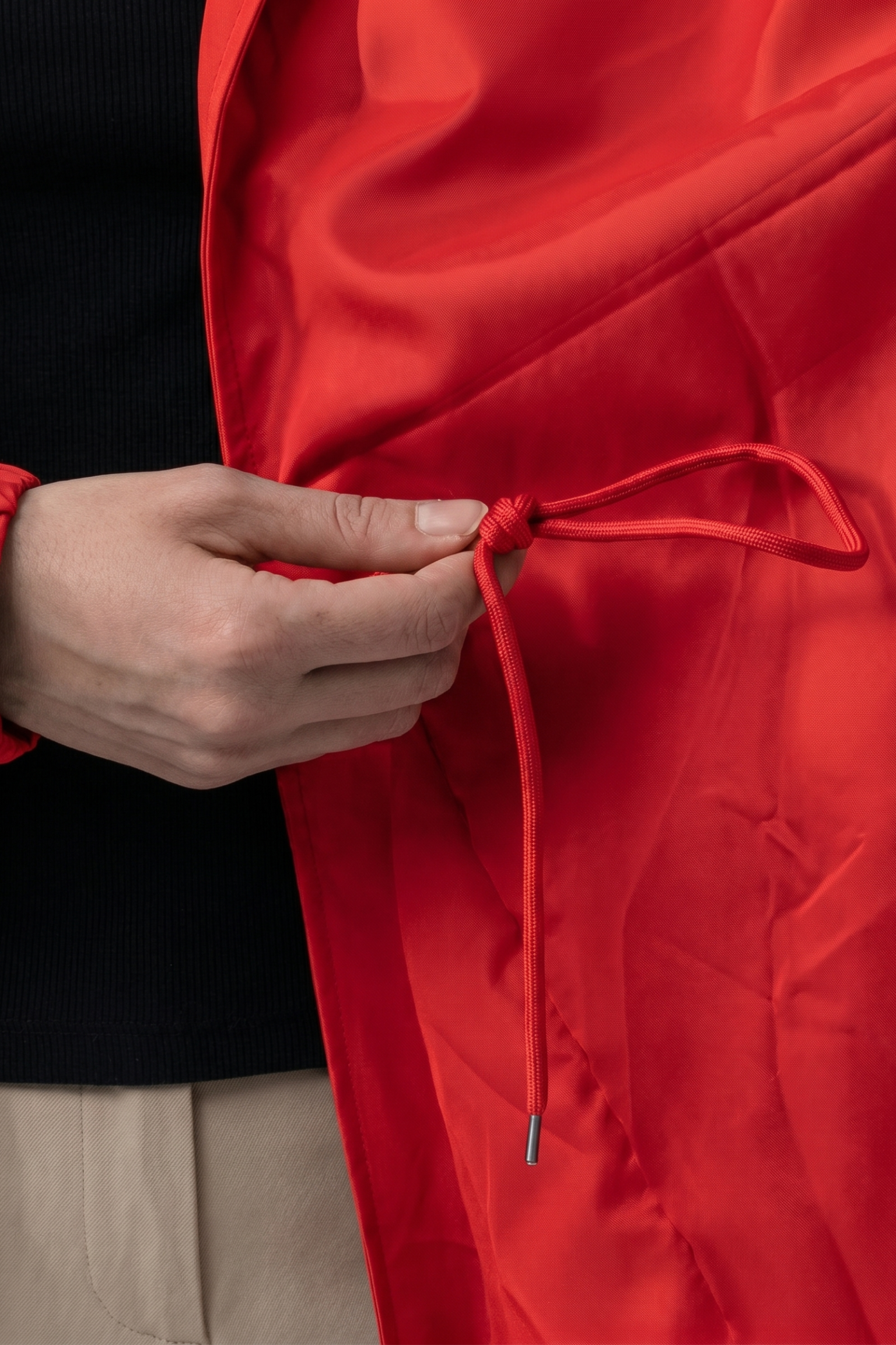 A person ties the inner red drawstring of The Water Resistant Hooded Oversized Windbreaker Rain Jacket while wearing a black top and beige pants.