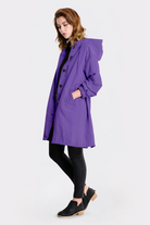 A person stands sideways wearing the knee-length Icon Windbreaker Rain Jacket in purple with large buttons and pockets, paired with black pants and low-heeled black slip-on shoes. The background is plain white.