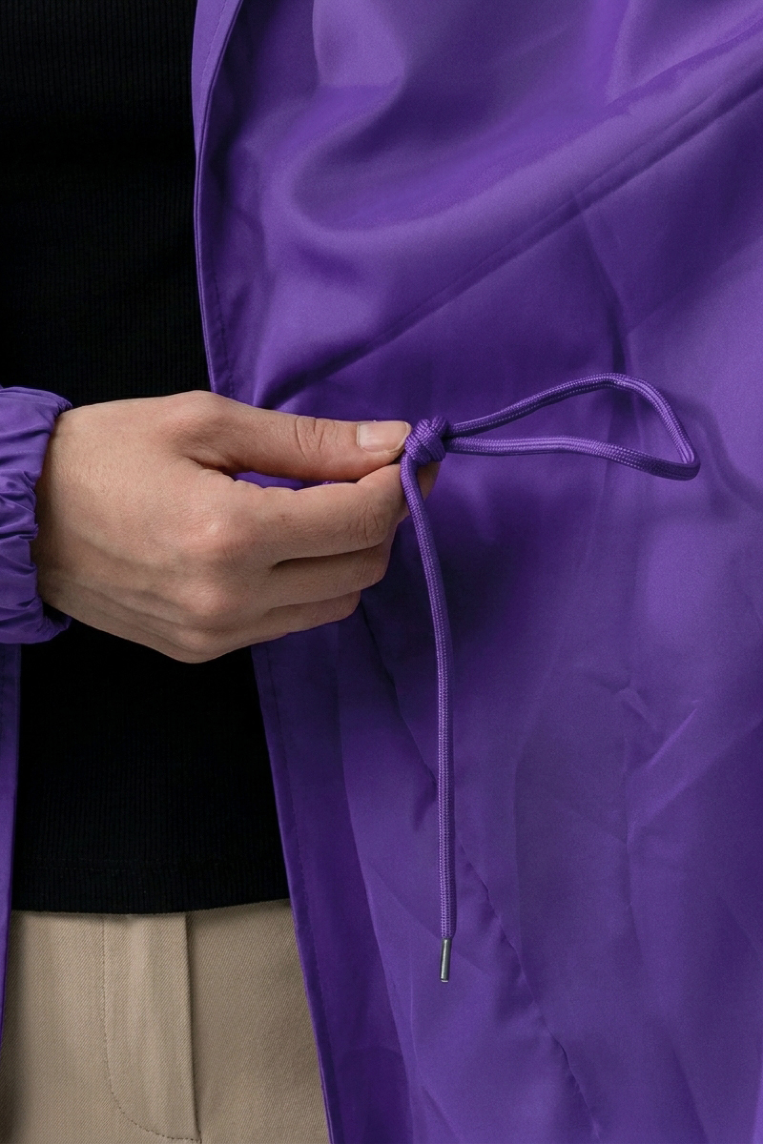 A person adjusts the drawstring of The Water Resistant Hooded Oversized Windbreaker Rain Jacket in purple, with only their hand, part of the jacket, and a portion of their beige pants visible.