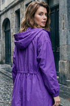 A woman with wavy brown hair stands on a cobblestone street, wearing the Icon Windbreaker Rain Jacket in purple. She looks over her shoulder toward the camera, with historic stone buildings in the background.