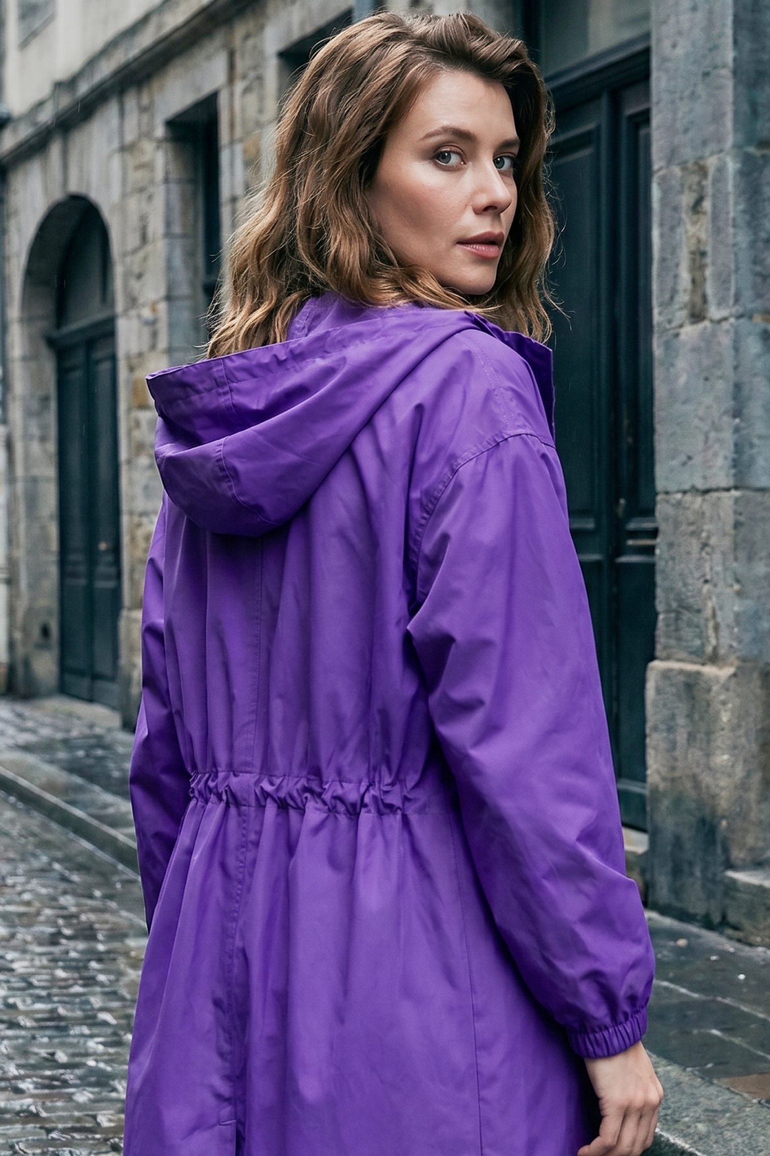 A woman with wavy brown hair wearing The Water Resistant Hooded Oversized Windbreaker Rain Jacket in purple stands on a cobblestone street beside stone buildings, looking back over her shoulder.