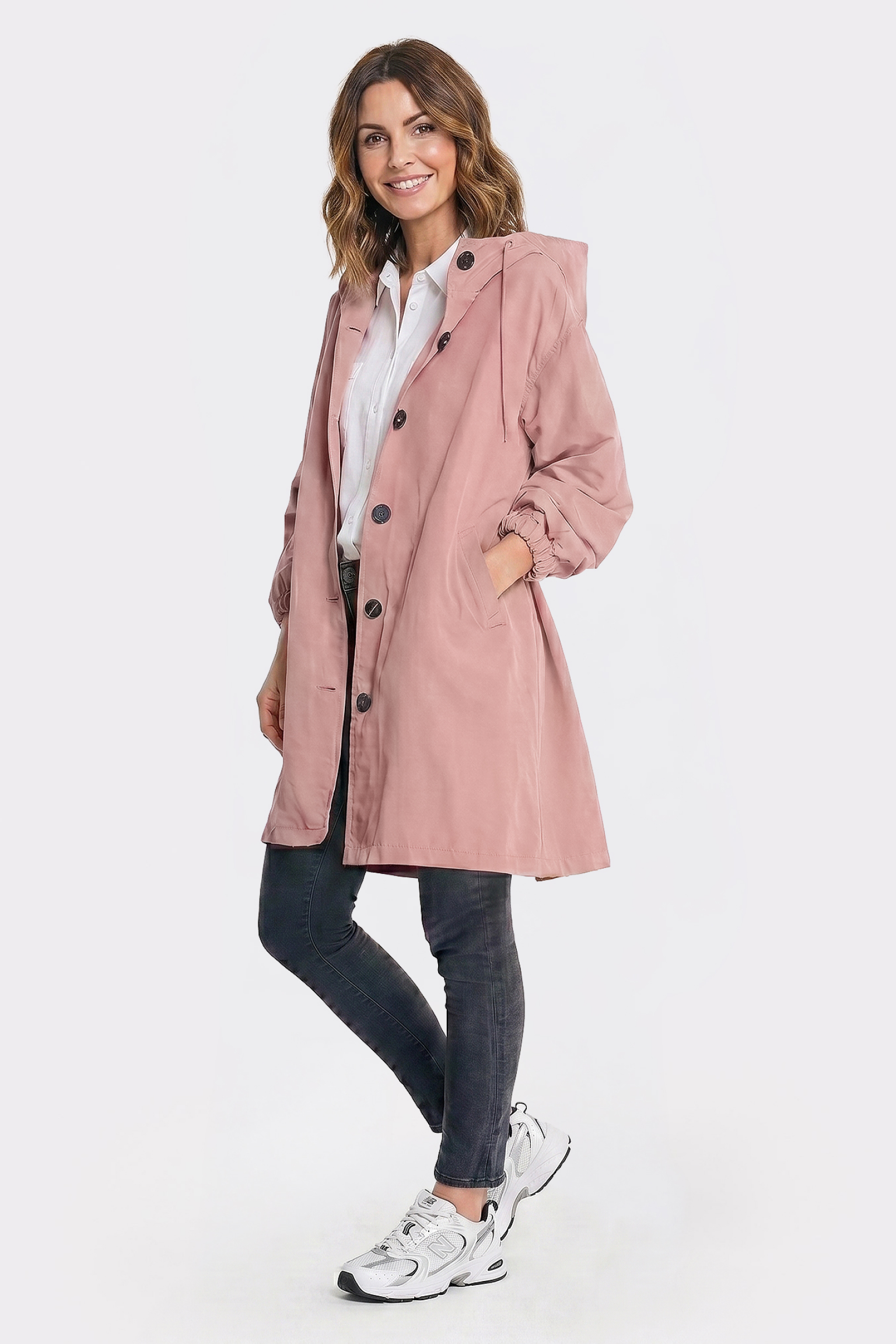 A woman with shoulder-length brown hair smiles as she faces forward, wearing the Icon Windbreaker Rain Jacket in light pink over a white shirt, paired with dark pants and white sneakers. One hand is in her jacket pocket. The background is plain white.