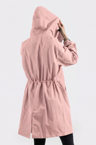 A person wearing the Icon Windbreaker Rain Jacket in light pink stands facing away from the camera, adjusting the hood. The background is plain and light-colored.