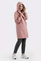A person models the Icon Windbreaker Rain Jacket in pink, paired with black jeans and white sneakers, standing against a plain light background and touching the hood while facing the camera.