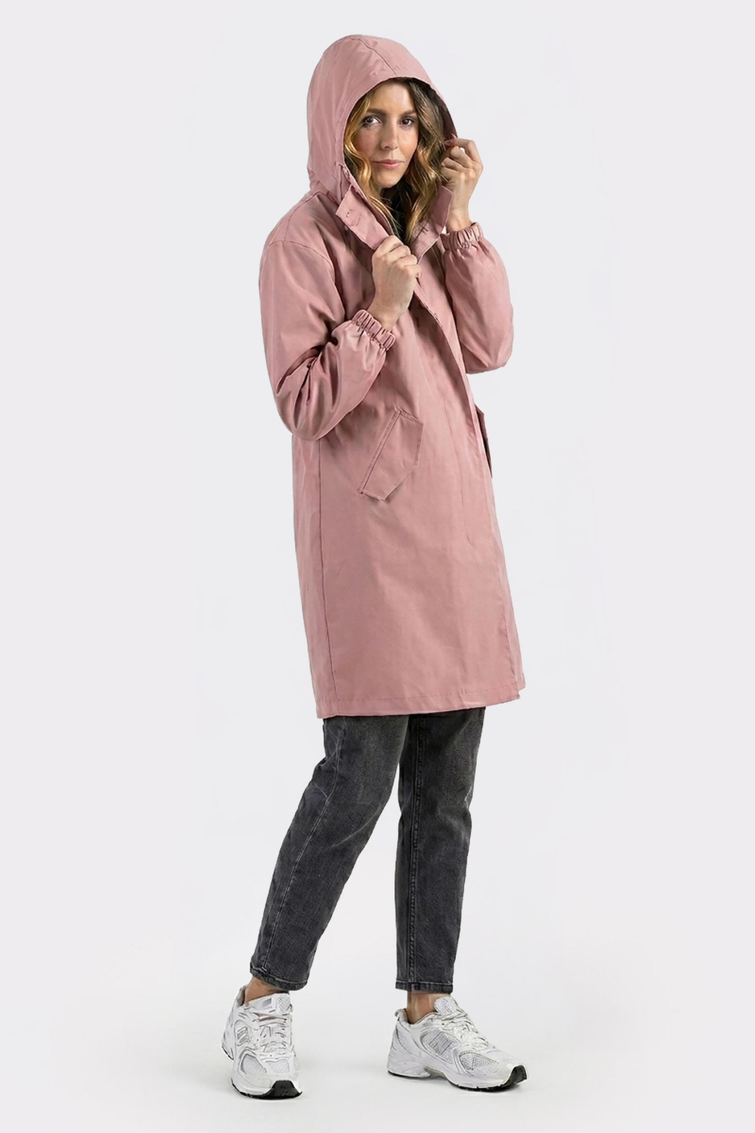 A person stands against a plain white background wearing The Water Resistant Hooded Oversized Windbreaker Rain Jacket in light pink, paired with dark jeans and white athletic shoes.
