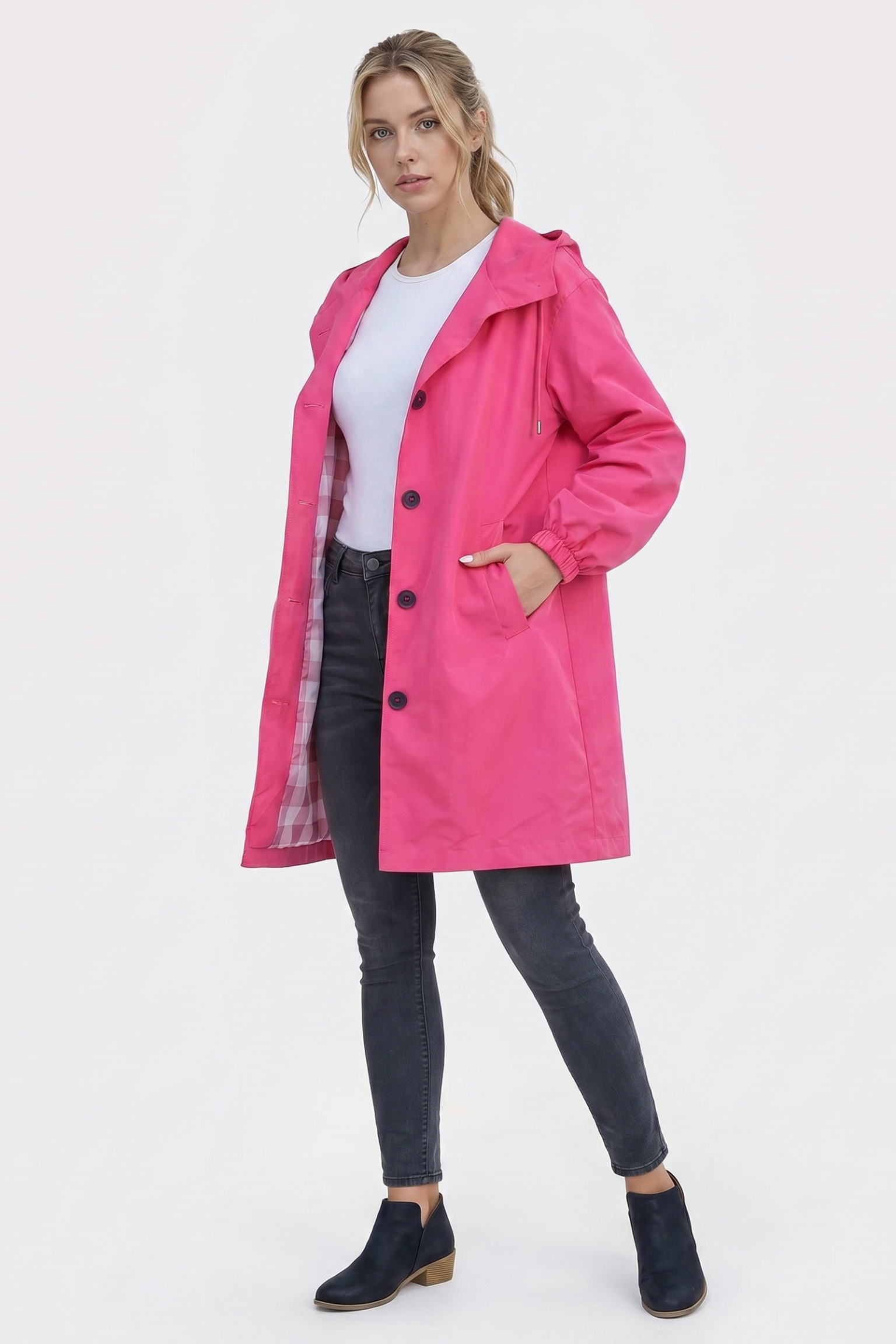 A woman stands against a plain white background wearing The Water Resistant Hooded Oversized Windbreaker Rain Jacket in bright pink, styled with black buttons, a white shirt, dark gray jeans, and black ankle boots.