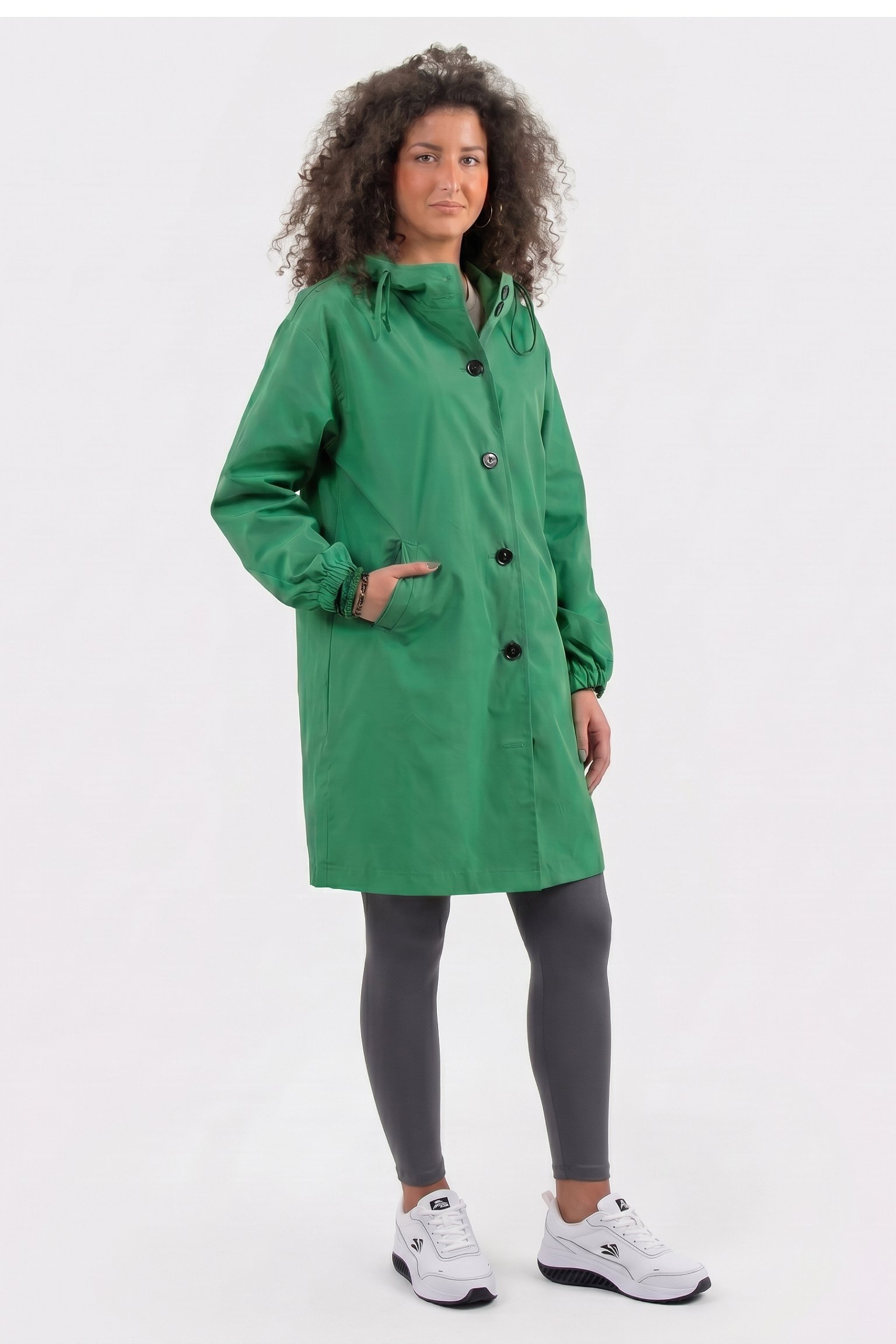 A person faces forward against a white background, wearing The Water Resistant Hooded Oversized Windbreaker Rain Jacket in green, styled with gray leggings and white sneakers. One hand is in a pocket while the other rests by their side.