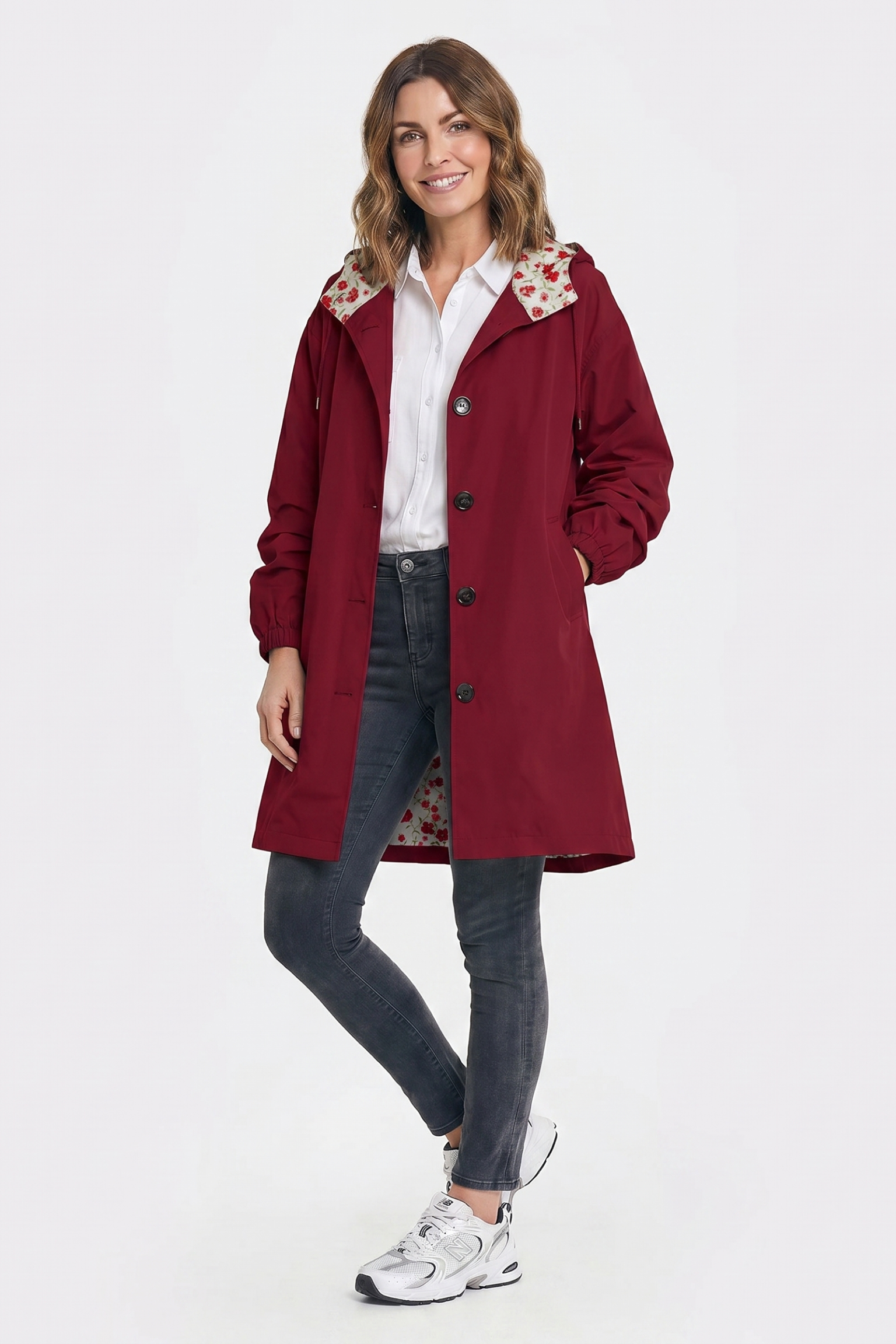 A woman smiles with one hand in her pocket, wearing The Water Resistant Hooded Oversized Windbreaker Rain Jacket in red over a white shirt, dark gray jeans, and white sneakers, standing against a plain white background.