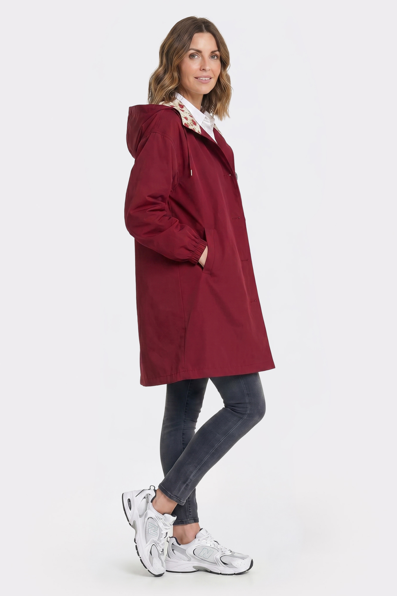 A woman in The Water Resistant Hooded Oversized Windbreaker Rain Jacket, dark skinny jeans, and white sneakers stands sideways against a plain white background, looking at the camera with a neutral expression. She has shoulder-length wavy hair.