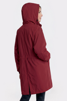 A person wears the Icon Windbreaker Rain Jacket, a long, hooded dark red coat with a straight fit and drawstring waist, standing slightly to the side with one hand in a pocket against a plain white background.