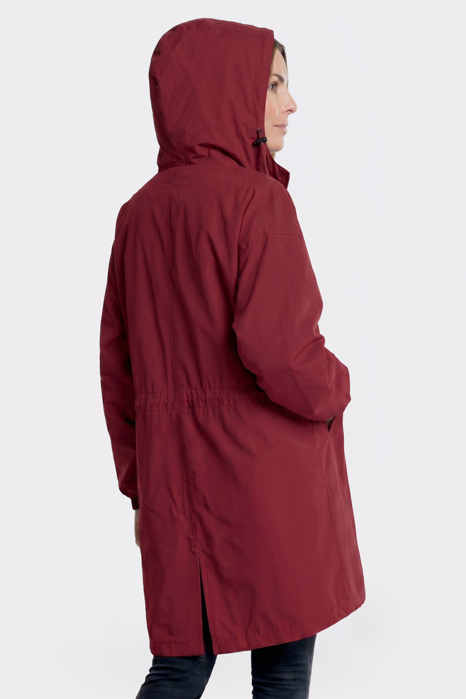 A person wears the Icon Windbreaker Rain Jacket, a long, hooded dark red coat with a straight fit and drawstring waist, standing slightly to the side with one hand in a pocket against a plain white background.