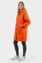 A person stands slightly sideways in an Icon Windbreaker Rain Jacket (orange), paired with black leggings and white sneakers, one hand in the coat pocket, set against a plain light background.