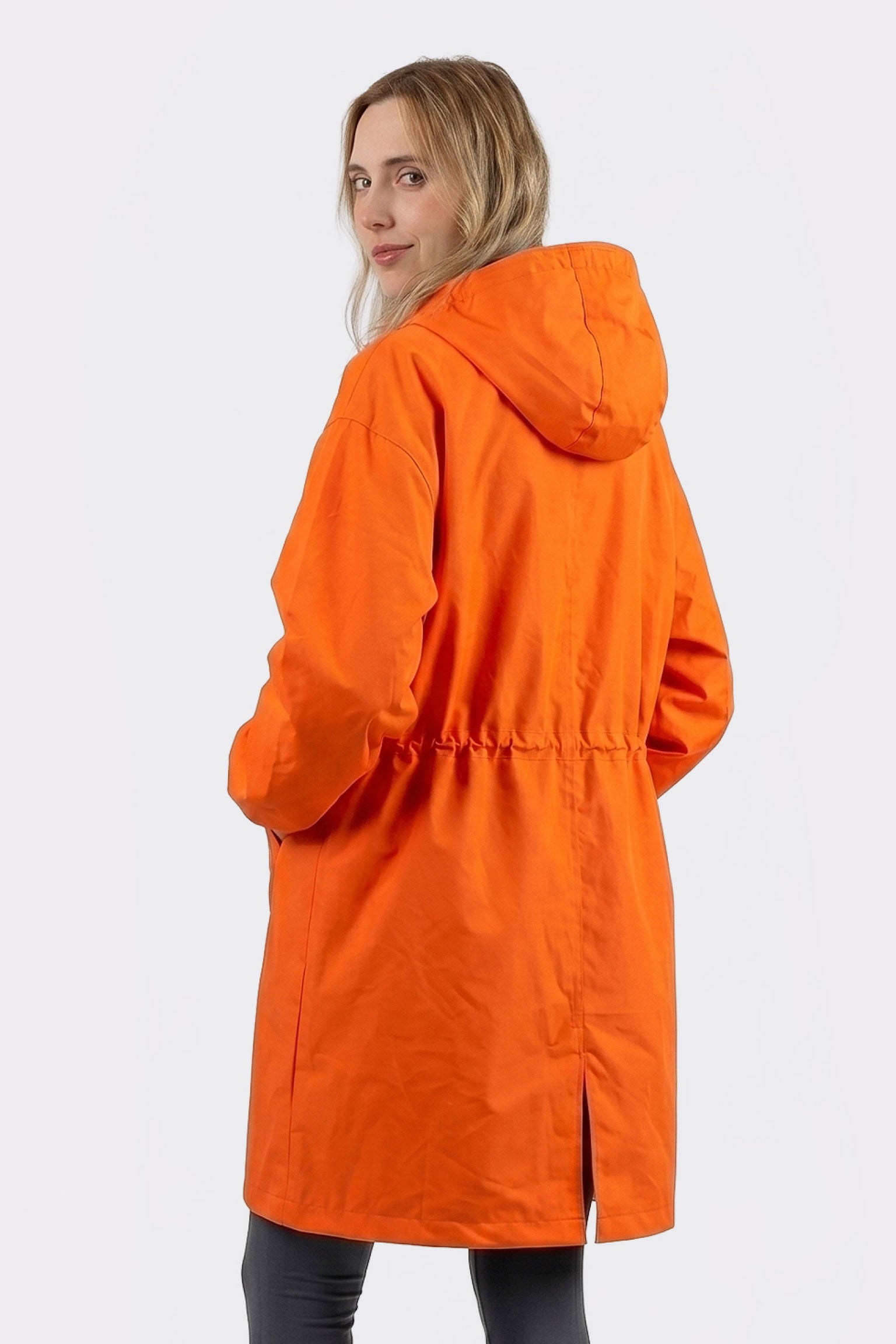 A person with long blonde hair is wearing The Water Resistant Hooded Oversized Windbreaker Rain Jacket in orange with a drawstring waist and back slit, paired with black pants, facing away and looking over their shoulder.