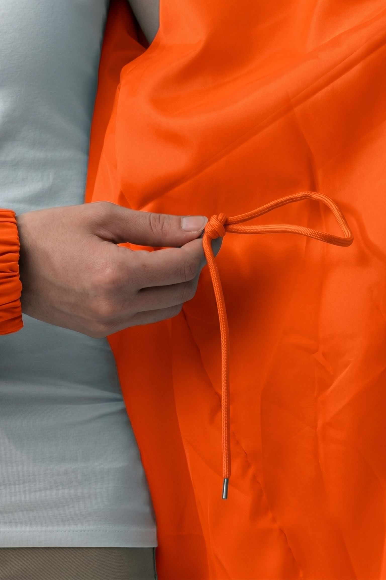 A person wearing a white shirt holds the drawstring of the Icon Windbreaker Rain Jacket in orange, with an orange scrunchie on their wrist.