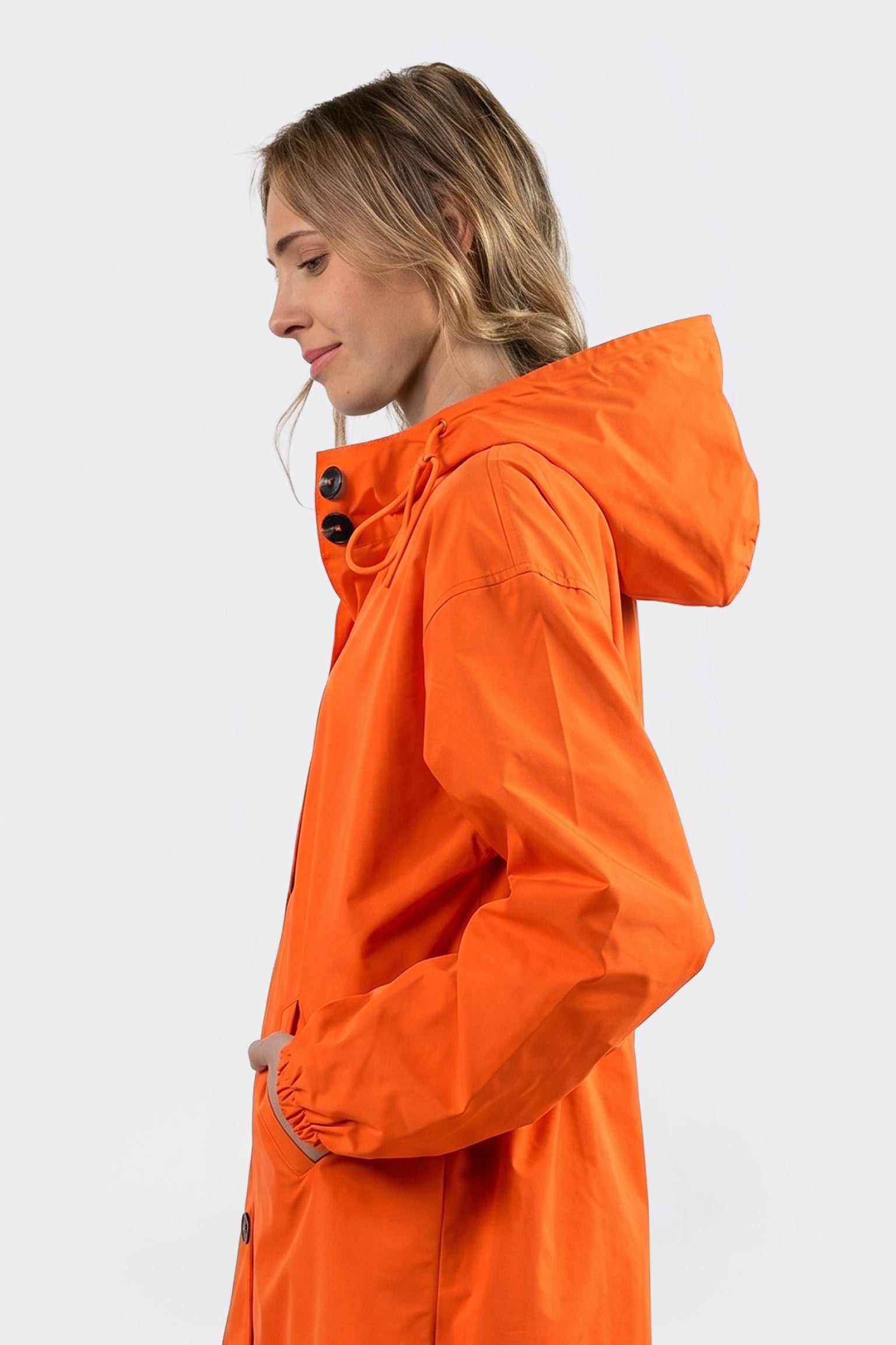 A person with light brown hair wears The Water Resistant Hooded Oversized Windbreaker Rain Jacket in orange, shown in profile against a plain, light background. One hand is in the pocket; black button details accent the hood.