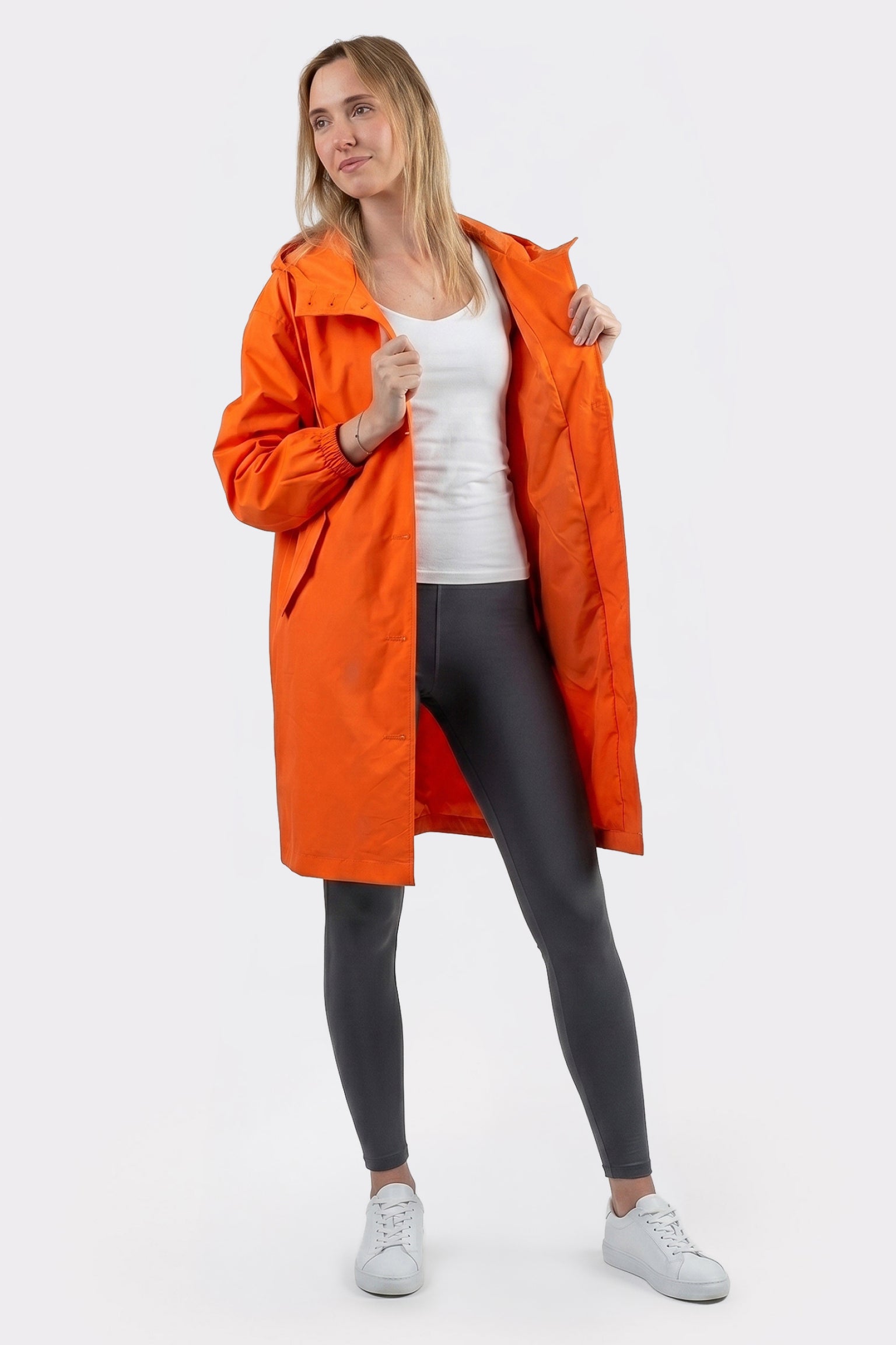 A person models The Water Resistant Hooded Oversized Windbreaker Rain Jacket in orange over a white shirt and black leggings, standing against a white background with the coat open, gazing slightly to the side.