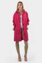 A woman faces forward in the Icon Windbreaker Rain Jacket, a bright pink knee-length coat, layered over a white top and beige pants with nude open-toed heels. She holds the sides of the unbuttoned jacket against a plain white background.